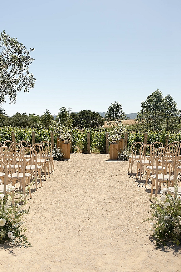 Ceremony setup with outdoor wedding ceremony aisle, wooden chairs and white-green florals on pedestals along a gravel path in vineyards