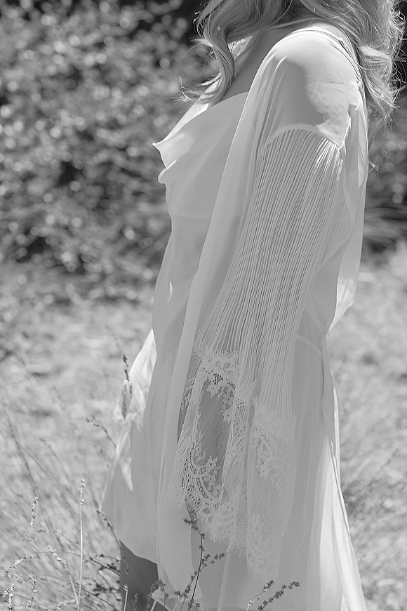 Bridal getting ready portrait in a lace bridal robe with pleated sleeves, hair in soft waves, standing in sunlit greenery outdoors