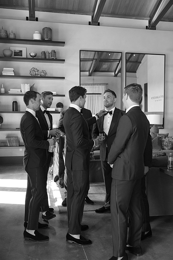 Groom getting ready as groomsmen getting ready help adjust his bow tie in a high-ceiling living room with exposed beams and mirrors
