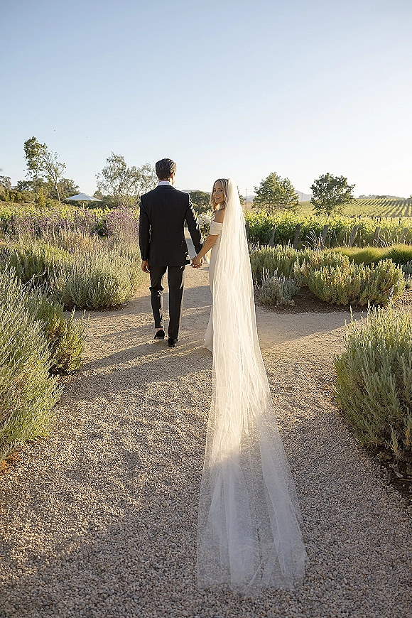 Couple portrait of newlyweds holding hands walking down a vineyard path, bride in strapless dress with veil and bouquet, looking back