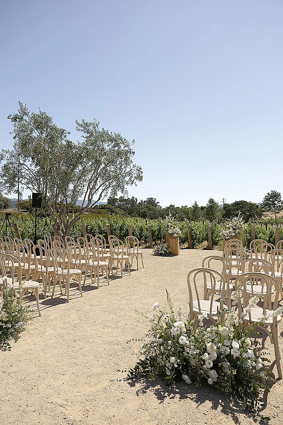 Ceremony setup with bentwood chairs and white floral arrangements lining a gravel aisle in a vineyard under blue sky with hills