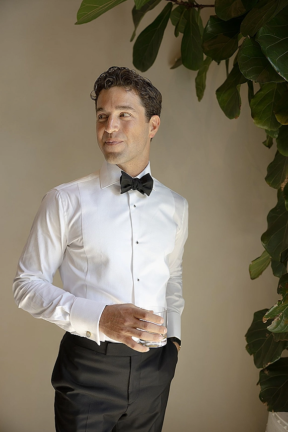 Groom portrait wearing a groom bow tie with crisp white tuxedo shirt, holding a drinking glass beside a neutral wall and plant leaves