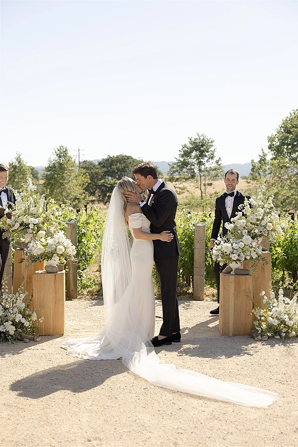 Wedding kiss as bride in off-shoulder gown and long veil holds groom’s face, outdoor ceremony kiss on vineyard aisle with white florals