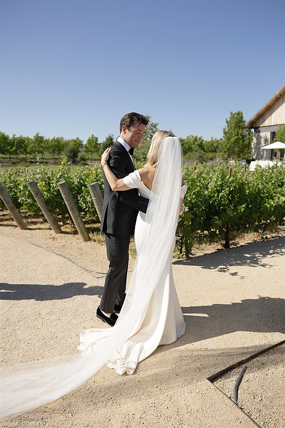 Couple portrait of bride and groom embracing, her wedding veil trailing behind on a vineyard gravel path near a barn under blue sky