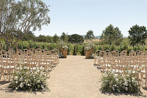 Ceremony setup with outdoor wedding ceremony chairs and white flower arrangements lining a gravel aisle, vineyard rows and hills beyond
