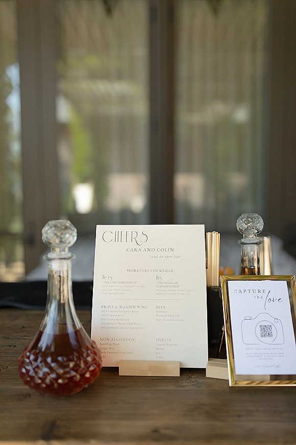 Wedding bar signage with signature cocktails sign and framed QR code on a wood counter, glass decanters and greenery bokeh behind