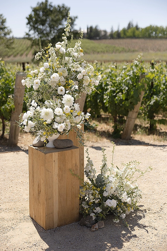 Ceremony floral arrangement in a ceramic vase with white and yellow blooms and greenery on a wooden pedestal along vineyard rows