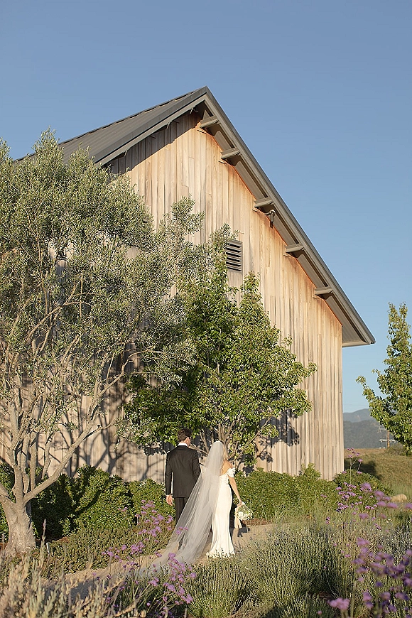 Couple portrait of bride and groom walking away, her long veil trailing and bouquet in hand, past a rustic barn garden with purple flowers