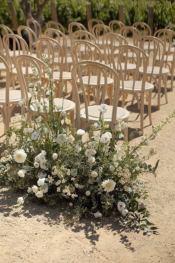 Ceremony seating with bentwood chair rows and white aisle flowers with greenery on a sunlit gravel garden aisle by hedges