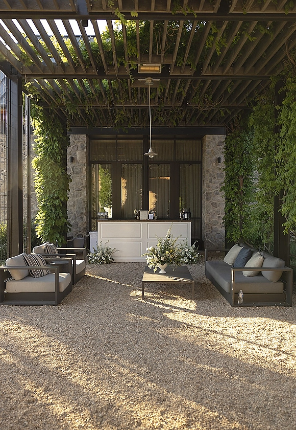 Wedding lounge area with outdoor sofas and throw pillows around a coffee table, with a bar and pendant light under a vine-covered pergola