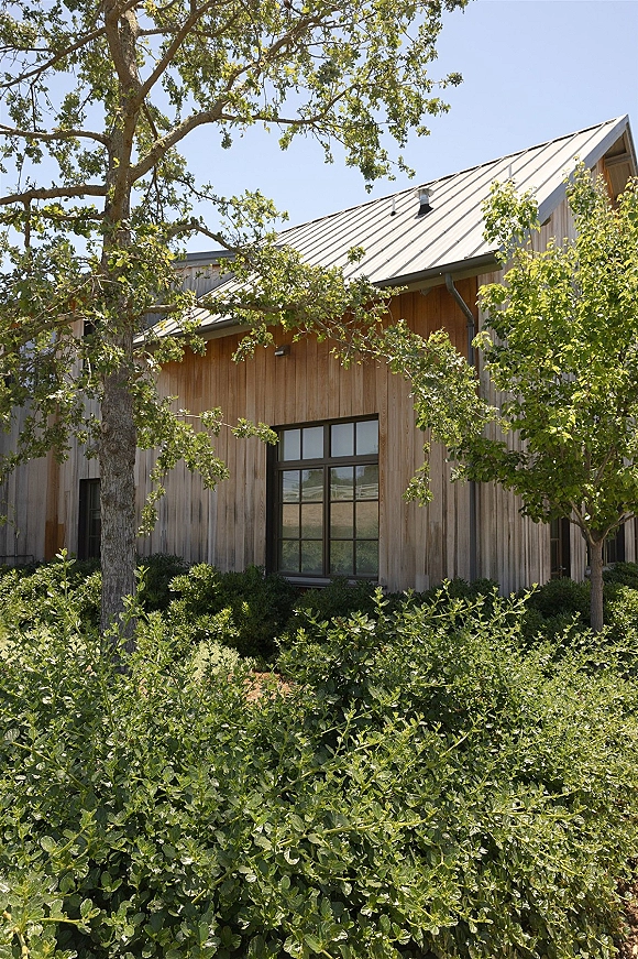 Wedding venue exterior with wood siding and black framed windows beneath a metal roof, surrounded by shrubs and trees under blue sky