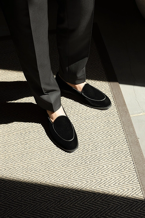 Groom shoes in black velvet loafers beneath dress pants, resting on patterned carpet with soft window light and floor shadows