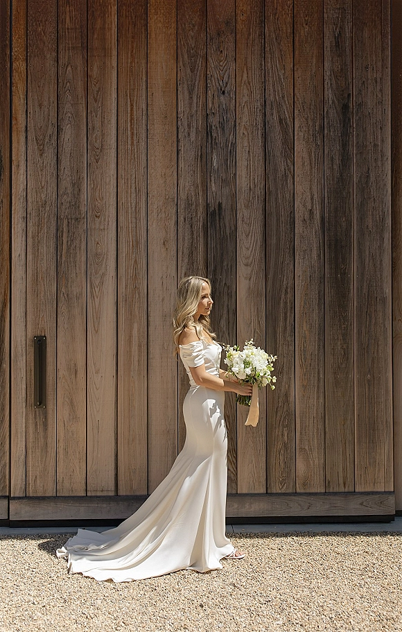 Bridal portrait of a bride in an off the shoulder wedding dress holding a white floral bouquet, long train flowing by a rustic wood wall in sunlight