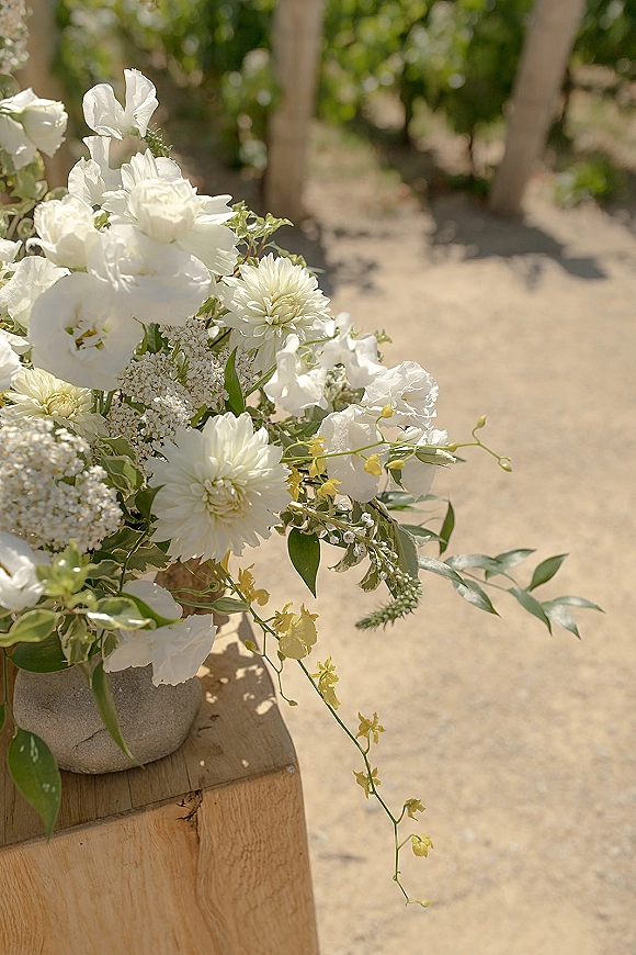 Wedding floral arrangement of white wedding flowers and greenery in a stone vase on a wooden pedestal along a sunlit vineyard path