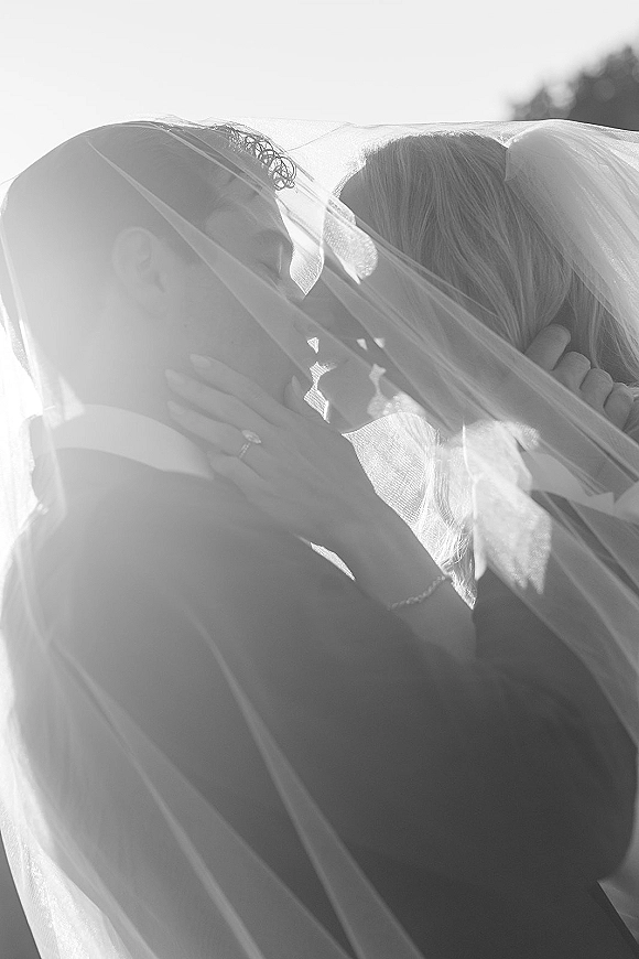 Wedding kiss portrait of bride and groom kissing under the veil, her ringed hand on his face, with sky and distant trees behind