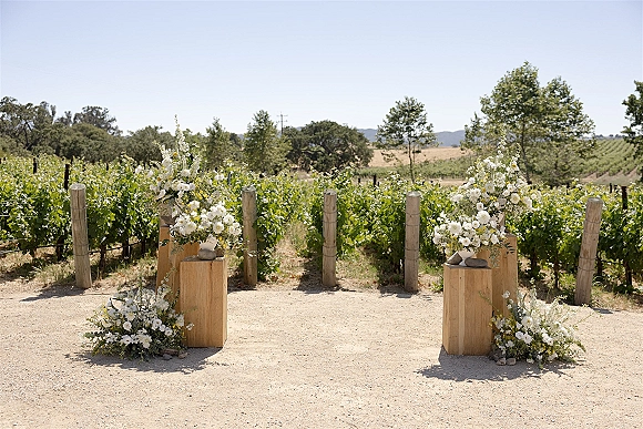 Ceremony aisle decor with outdoor ceremony aisle florals on wooden pedestals and stone vases, lining a dirt path between vineyard rows