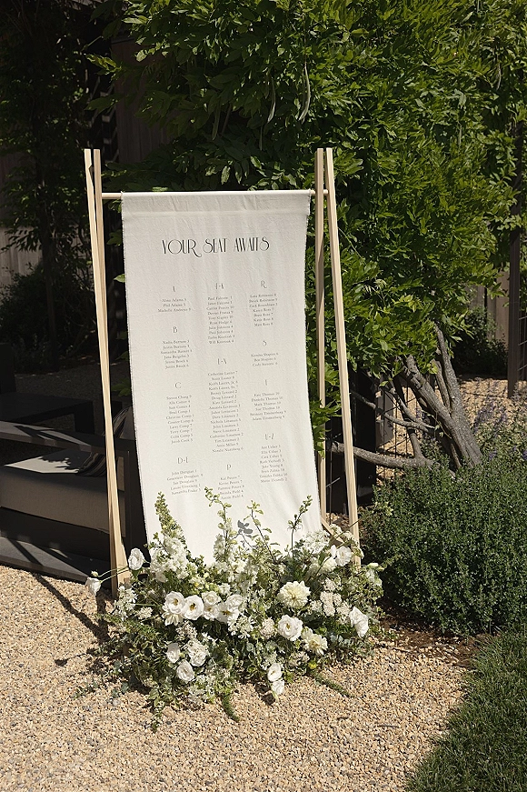 Wedding seating chart on a linen seating chart sign displayed on a wooden stand with white flowers and greenery in an outdoor garden setting