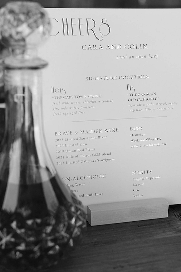 Wedding bar menu printed in a stand on a wood tabletop beside a glass decanter and bottle, with blurred glassware in foreground