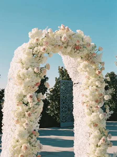 Wedding floral arch with asymmetrical round design of white flowers, blush roses, and hydrangeas beside a decorative screen on an outdoor patio