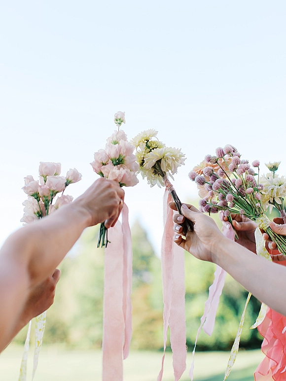 Bridesmaid bouquets held up in hands, showing bridesmaid bouquet ideas with pink ribbon streamers against a bright sky and greenery backdrop