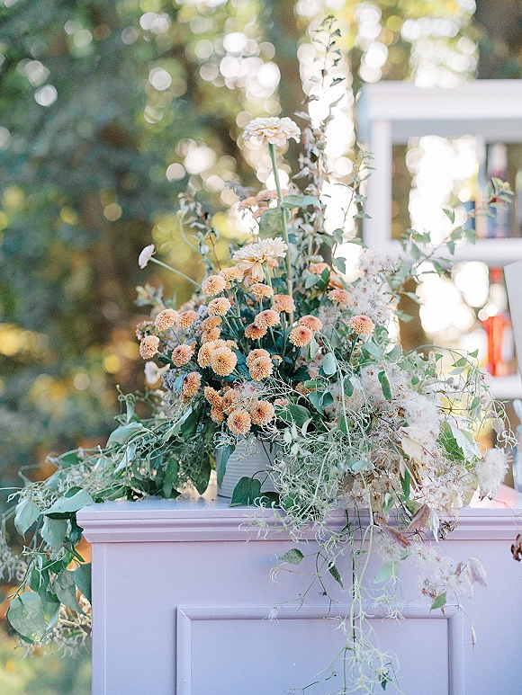 Wedding floral arrangement with ceremony pedestal flowers, trailing vines and dried florals on a white pedestal in sunlit garden trees