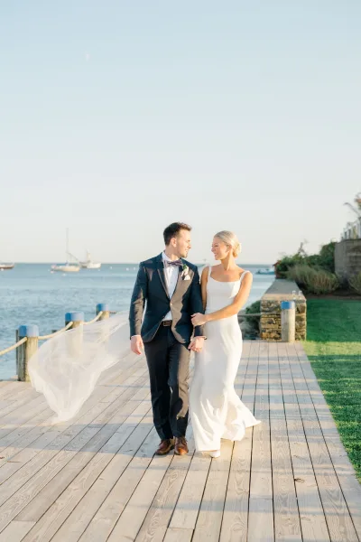 Couple portrait of bride and groom walking arm in arm as her veil blows, with ocean and sailboats behind on a wooden boardwalk