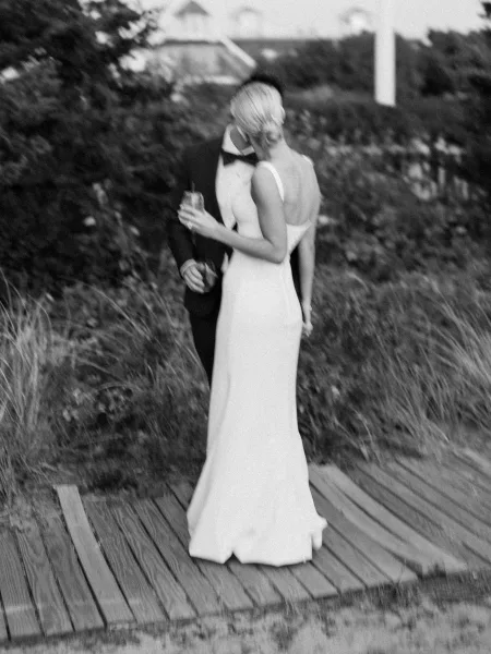 Wedding kiss portrait of bride and groom kissing, her backless dress and updo, his tuxedo and bow tie, holding drinks on a wooden walkway.