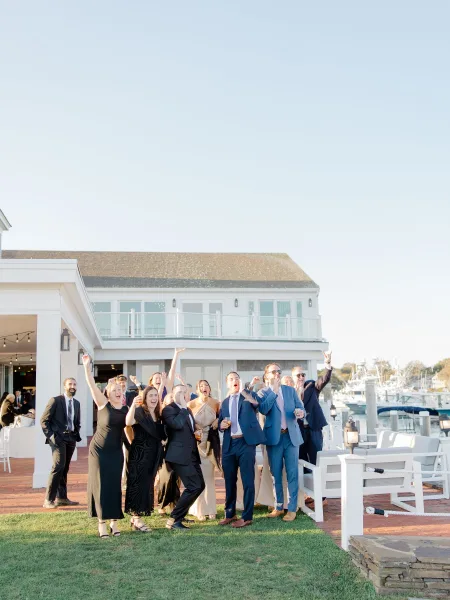 Wedding guest group photo of guests cheering with cocktail drinks by a waterfront marina, string lights overhead, boats and blue sky behind