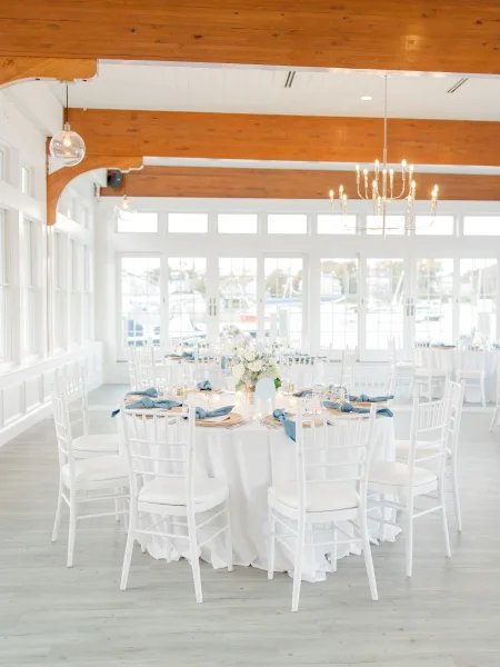 Reception tablescape with blue napkins and gold chargers on a round table, votive candles and white florals beneath chandeliers by marina-view windows