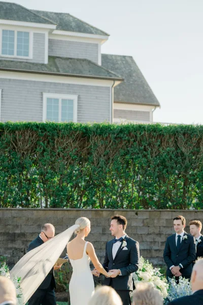 Wedding vows during an outdoor wedding ceremony as bride in long veil and groom in tuxedo hold hands before altar florals and guests
