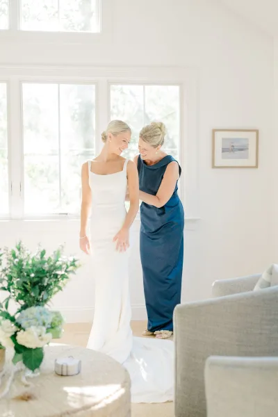 Bride getting ready as a bridesmaid helps with a wedding dress fitting, adjusting the train in a bright living room by large windows.