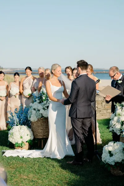 Ceremony moment with bride and groom holding hands during wedding vows, bridal party behind them on an oceanfront lawn by a stone wall