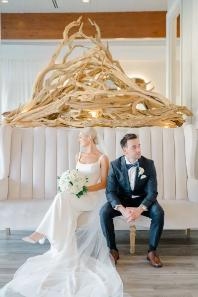 Couple portrait of bride and groom sitting on an upholstered sofa, her veil draped and white bouquet held beneath driftwood wall art
