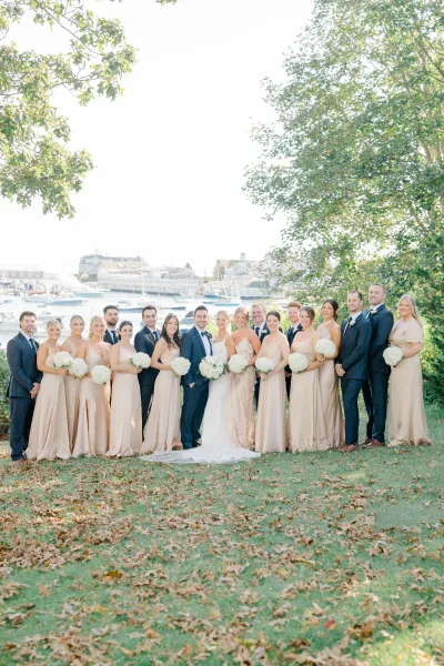 Wedding party portrait of bride and groom with bridesmaids in blush dresses and tuxedoed groomsmen on a harbor lawn with sailboats behind