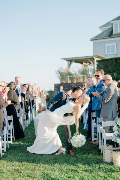 Wedding kiss moment as groom dips the bride in the aisle, her long veil trailing, guests cheering under a clear sky on the lawn