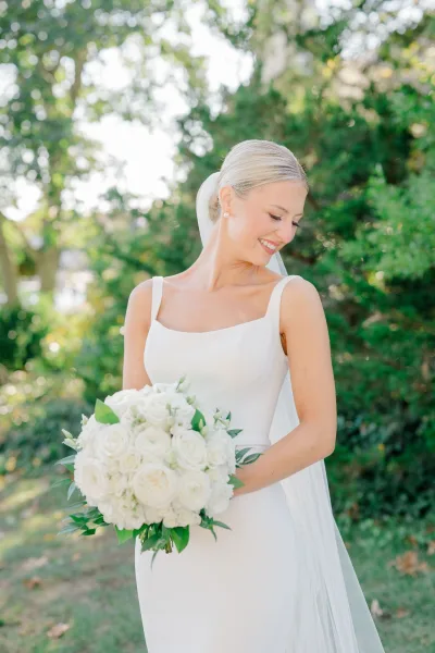 Bridal portrait of a bride holding a white rose bouquet in a minimalist satin wedding dress and long veil, looking down in sunlit greenery