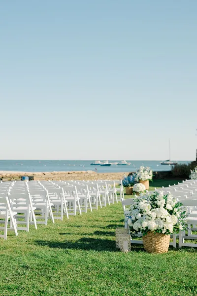Ceremony setup with white folding chairs and white rose floral baskets lining a grassy aisle, facing the ocean and rocky shoreline beyond