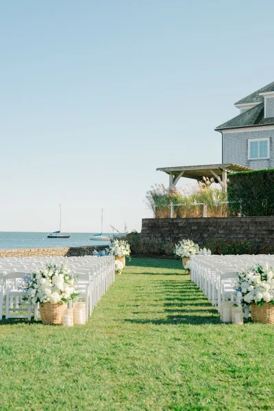Ceremony setup with white folding chairs lining a flowered aisle, wicker baskets and lanterns on a coastal lawn overlooking ocean sailboats