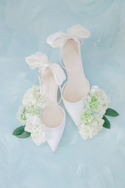 Bridal shoes with pearl-trim ankle straps and white bow heels, styled with hydrangea blooms on a blue textured backdrop flat lay