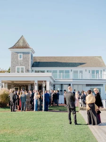 Wedding cocktail hour with wedding guests mingling around cocktail tables under string lights on a patio beside a shingle house with tower