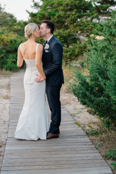 Wedding kiss portrait of bride and groom kissing on a wooden boardwalk by sand and evergreens, bride in strapless gown with pearl earrings