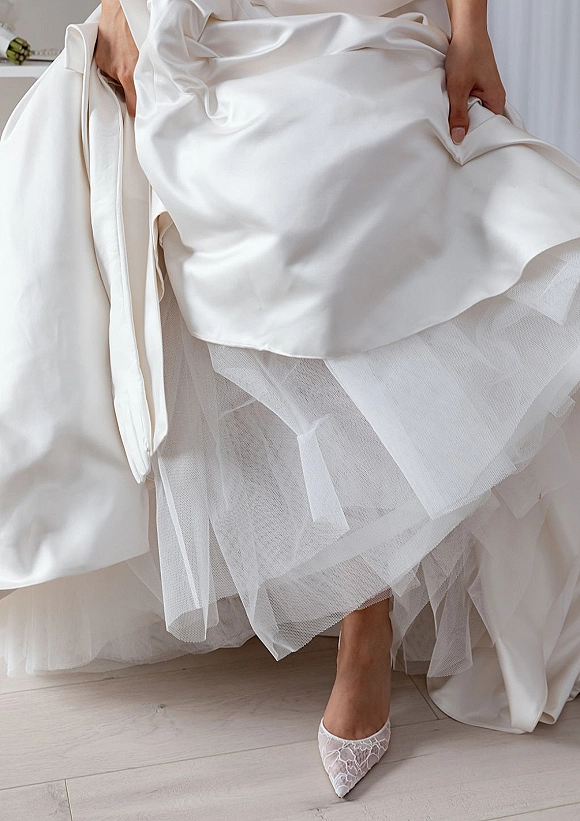 Wedding dress close-up showing a satin wedding dress hem over layered tulle underskirt beside lace bridal heels on a light wood floor