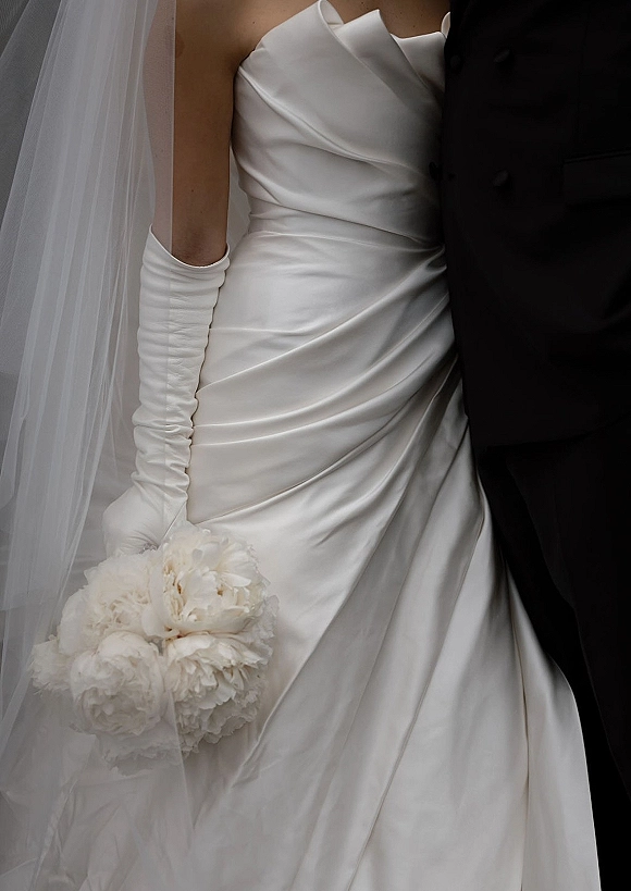 Bridal gown details in a strapless satin wedding dress with ruched bodice, opera gloves, veil, peony bouquet, and tux jacket on neutral backdrop