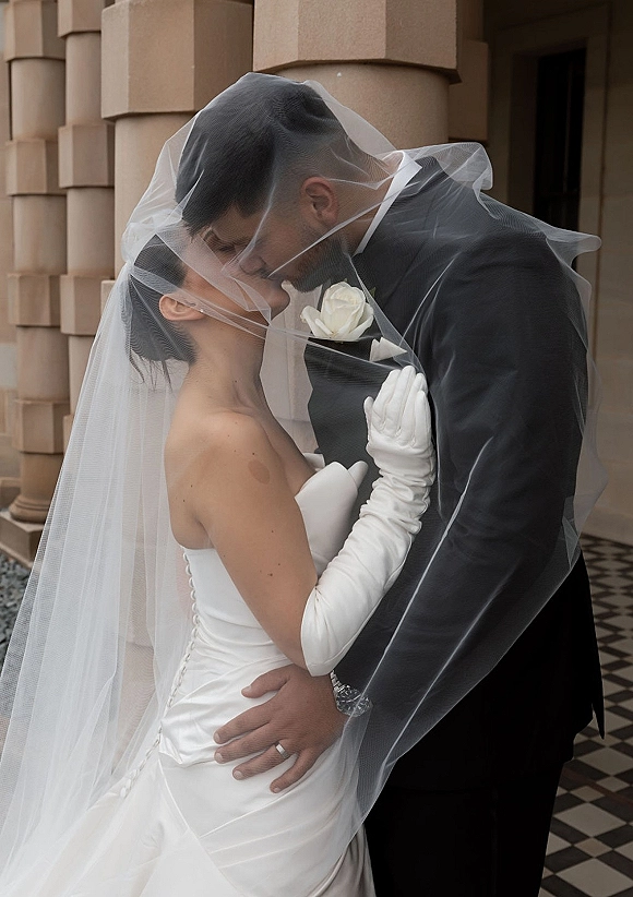 Wedding kiss portrait of bride and groom embracing, veil over face kiss with long gloves and tux, framed by stone columns entrance
