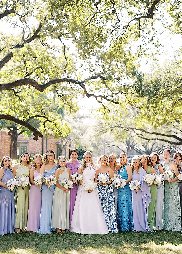 Bridesmaid group photo with bride and bridesmaids holding bouquets in mismatched dresses on a sunny lawn beneath oak trees by a brick building