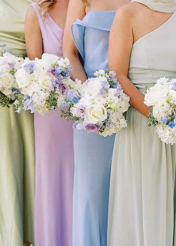 Bridesmaid bouquets with white roses, blue and lavender blooms, and greenery held against pastel bridesmaid dresses on a neutral backdrop