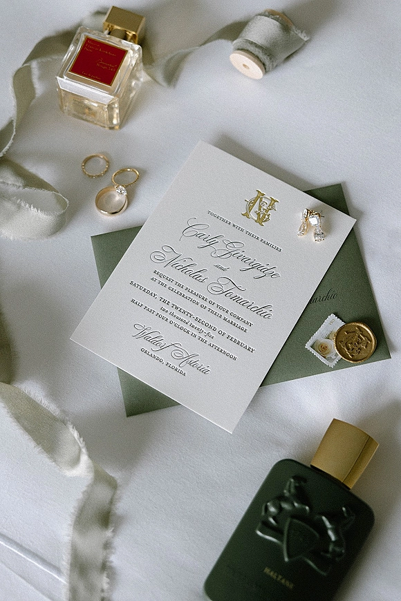 Wedding invitation suite with formal wedding invitation cards, sage green envelope, gold wax seal, rings, earrings and perfume on white fabric backdrop