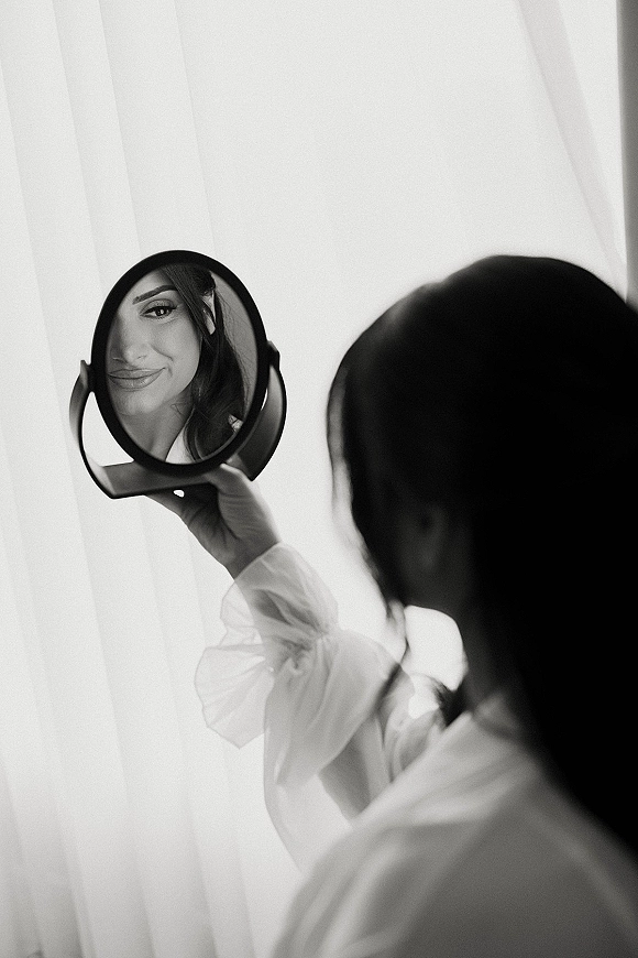 Bridal portrait of a bride in a robe holding a handheld mirror, softly lit by window light with sheer curtains behind her