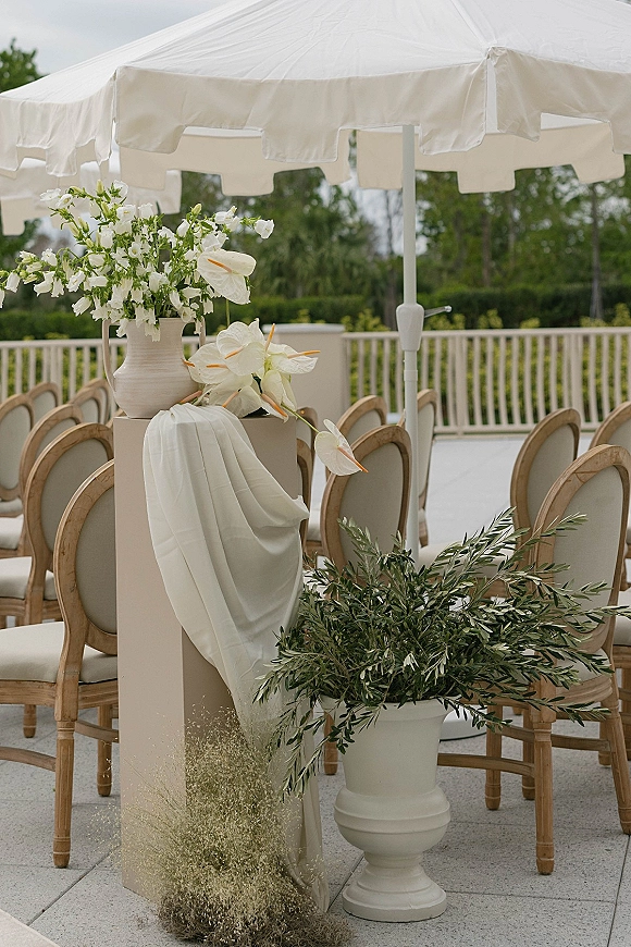 Ceremony aisle decor with outdoor ceremony chairs, white umbrella canopy, and a pedestal plinth of white anthuriums and greenery on a patio