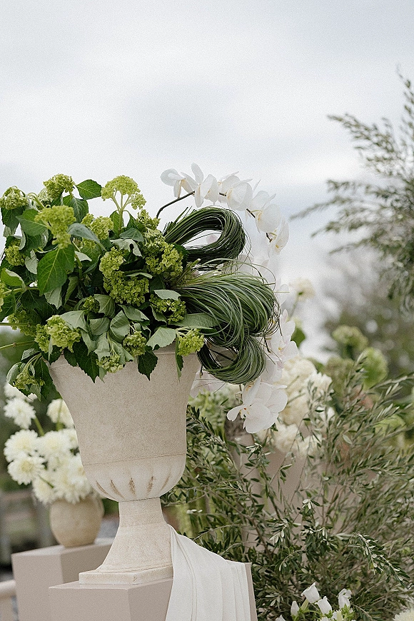 Ceremony floral arrangement in a stone urn with green hydrangea, white orchids, and greenery beside draped white fabric in a garden setting